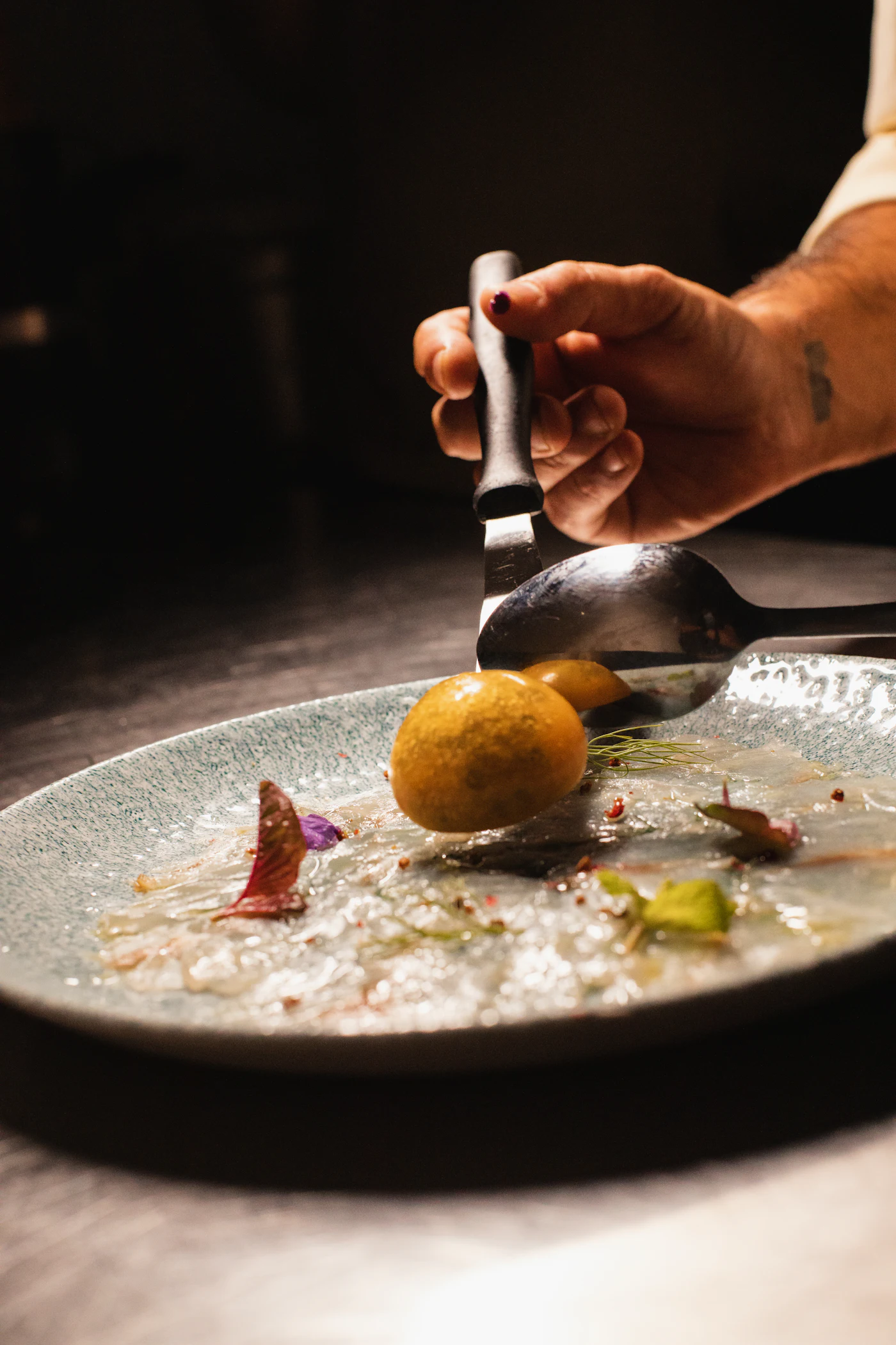 A chef spooning dressing over thinly sliced raw fish on a pale stoneware plate