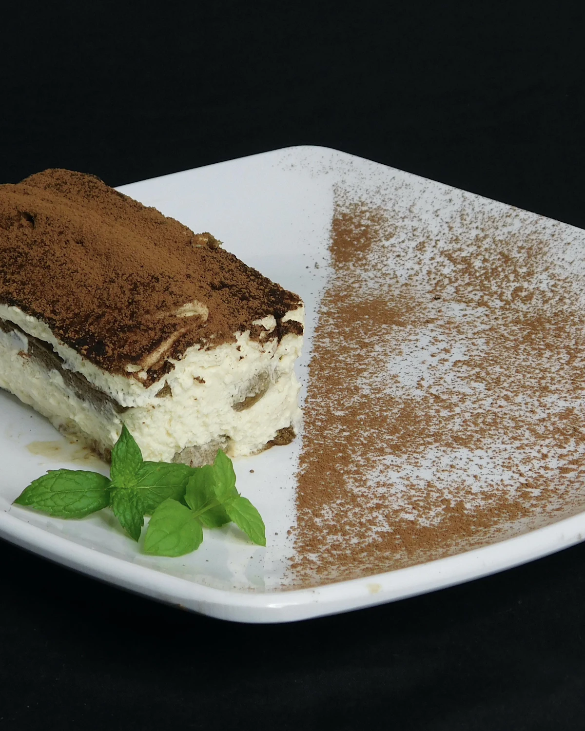 A slice of tiramis&ugrave; dusted with cocoa on a white plate against a black background