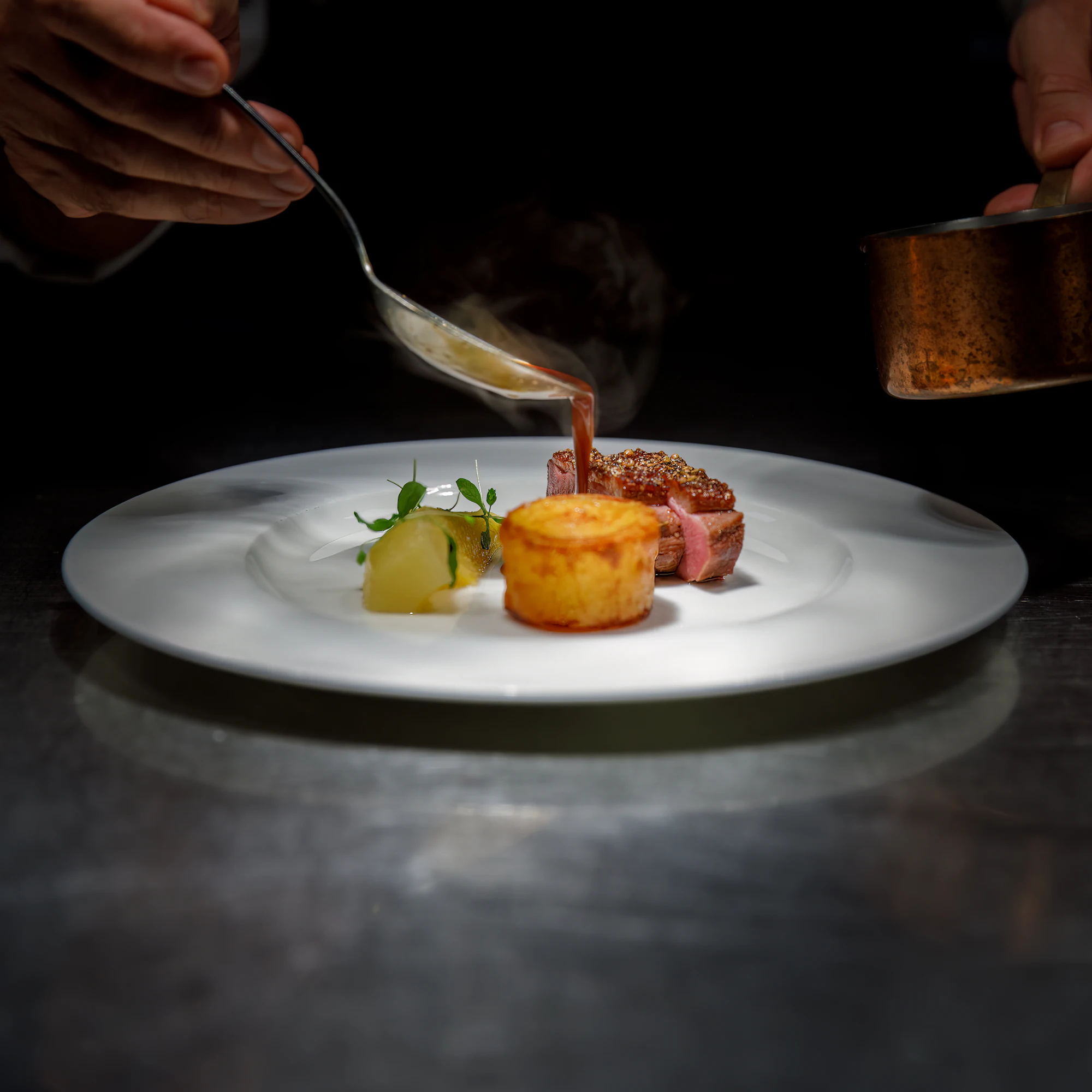 A chef spooning sauce onto a plated dish at a dark kitchen pass